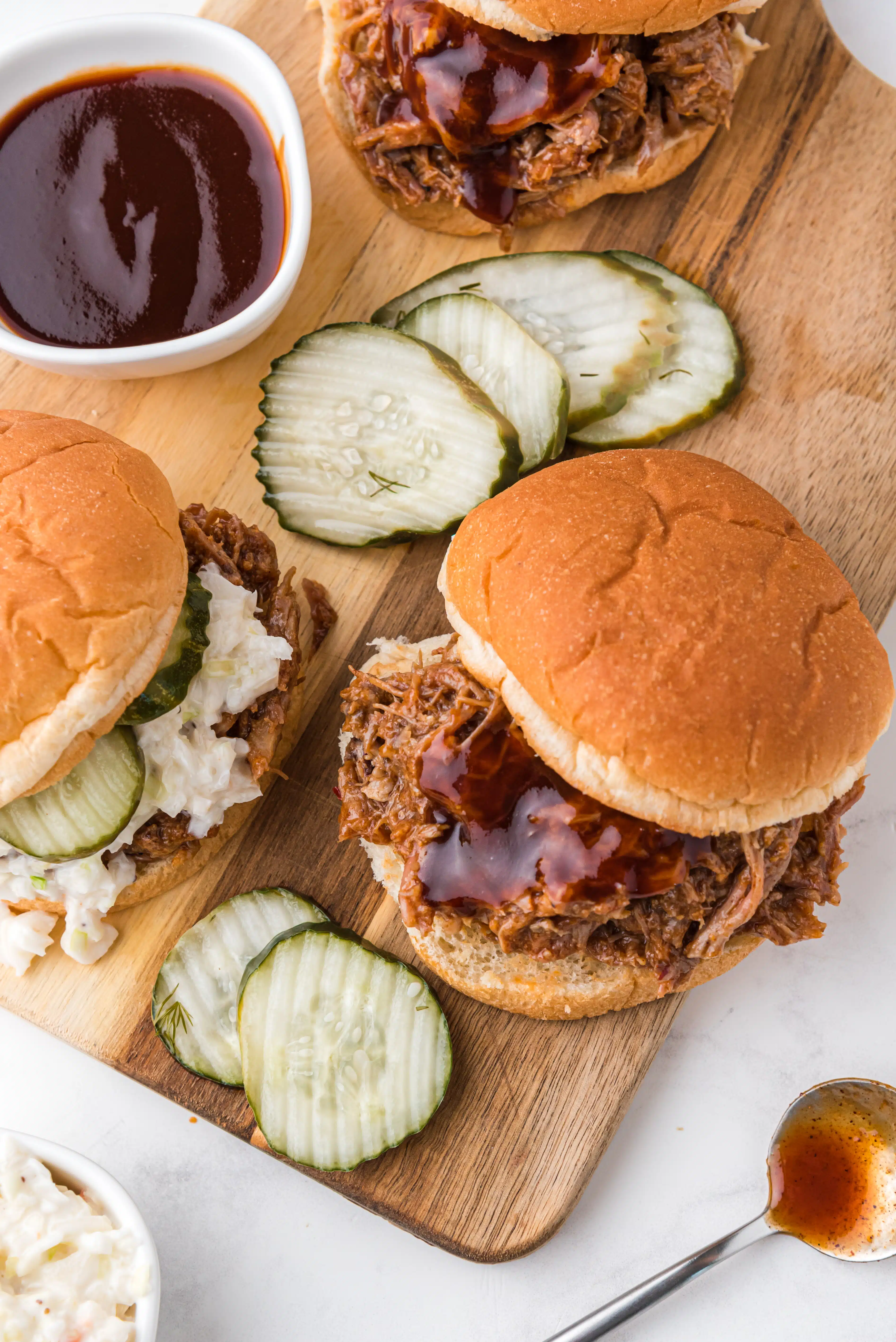 Small wood cutting board with two hamburger buns topped with bbq pulled pork and one topped with creamy coleslaw and pickles.