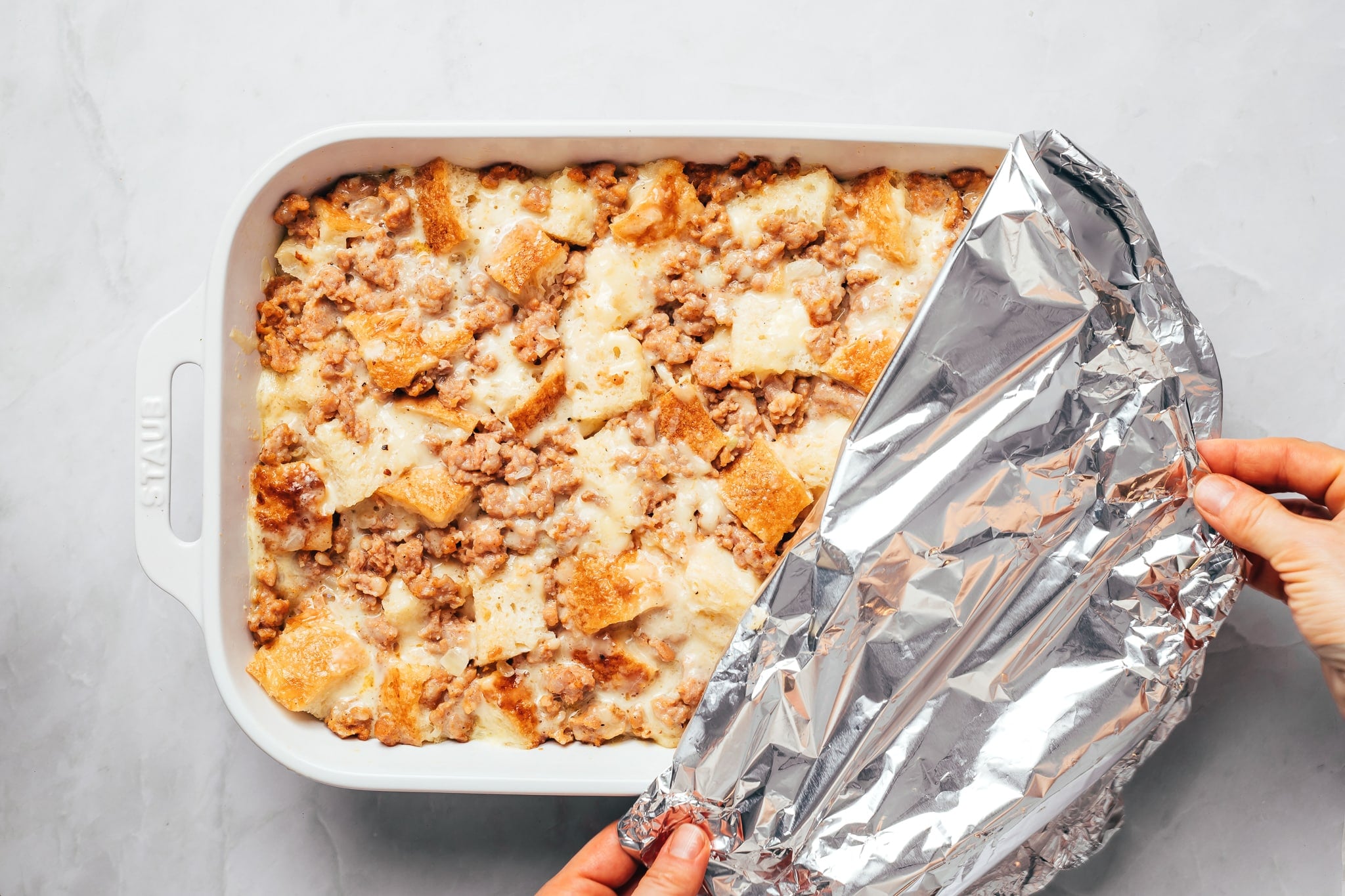 An unbaked sausage breakfast casserole in a white rectangular ceramic baking dish, filled with cubes of sourdough bread, crumbled sausage, shredded cheese, and a creamy egg mixture. Two hands are pulling back a large sheet of crinkled aluminum foil, revealing the assembled casserole underneath.