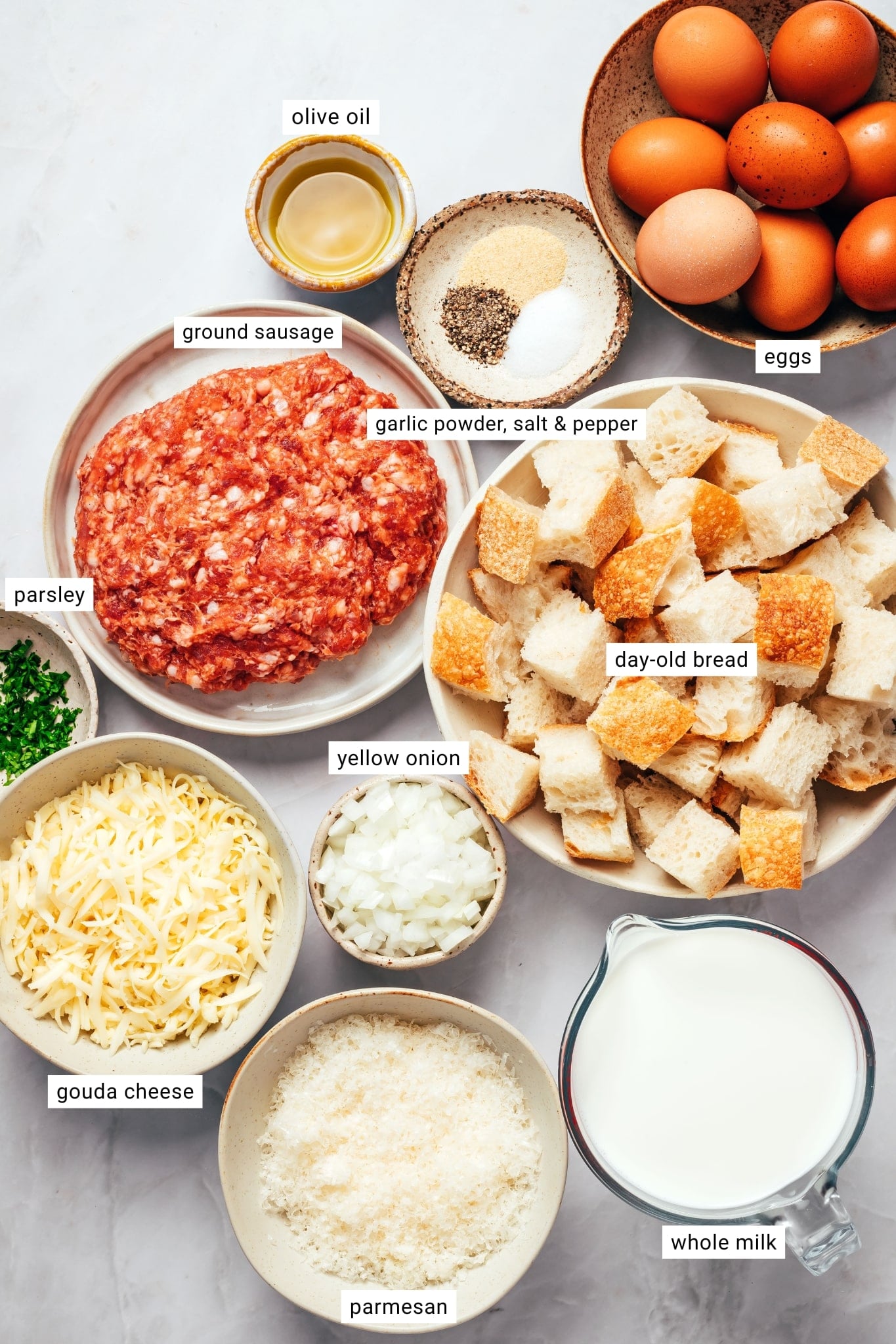 Ingredients for sausage breakfast casserole arranged in bowls and labeled with their names: raw ground sausage, brown eggs, cubed day-old sourdough bread, whole milk, shredded Gouda cheese, grated Parmesan, diced yellow onion, olive oil, garlic powder, salt, black pepper, and fresh chopped parsley.