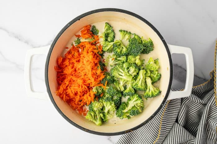 Overhead photo of bright green broccoli florets and a mound of shredded orange carrots added to a creamy soup base in a cream colored enameled Dutch oven with a black rim and white handles, with a black and white striped towel with yellow trim on the right.