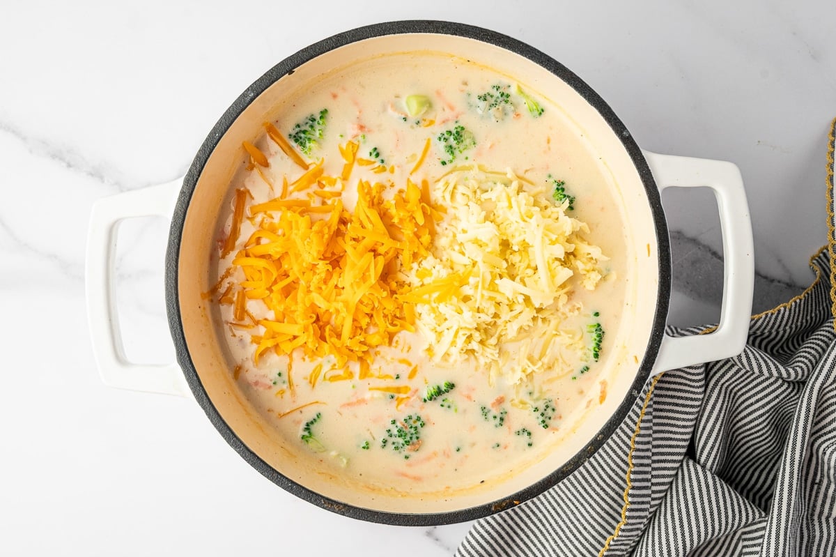 Overhead photo of shredded orange sharp cheddar and shredded white cheddar piled on top of creamy broccoli soup in a cream colored enameled Dutch oven with a black rim and white handles, with broccoli pieces visible around the edges and a black and white striped towel with yellow trim on the right.