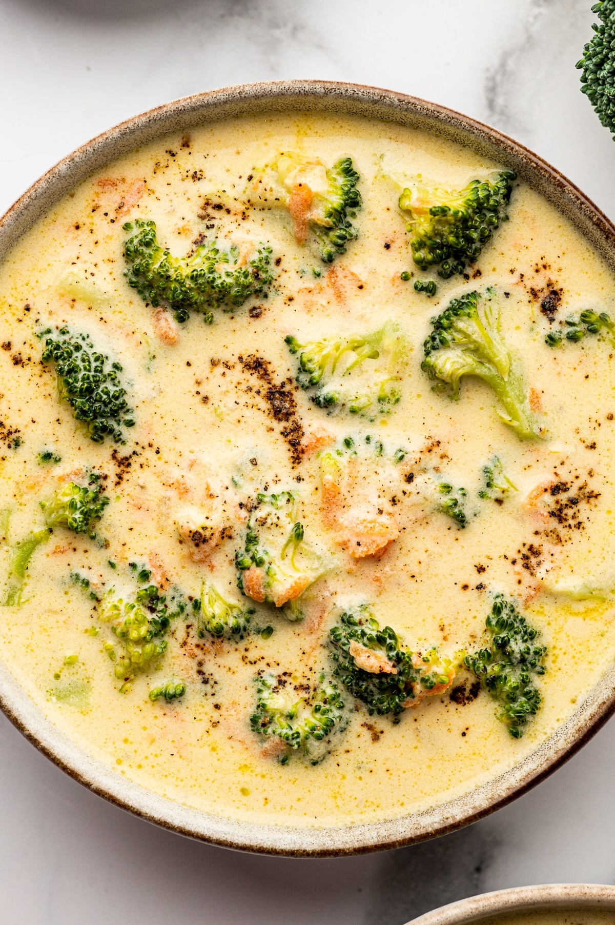 Close up photo of a beige speckled bowl filled with creamy broccoli cheddar soup with bright green broccoli florets and small carrot bits, topped with scattered cracked black pepper on the surface.