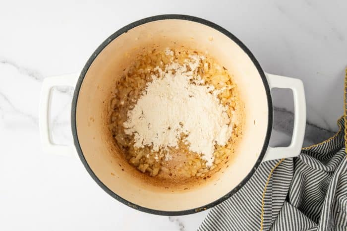 Overhead photo of a mound of all purpose flour sprinkled over softened chopped onions in a cream colored enameled Dutch oven with a black rim and white handles, with light browning on the sides of the pot and a black and white striped towel with yellow trim on the right.