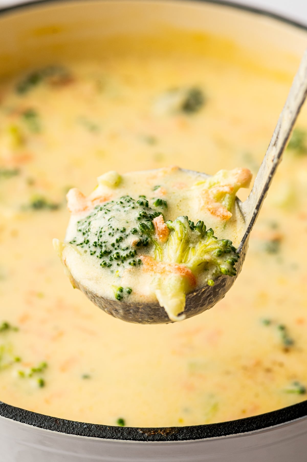 Close up photo of a metal ladle holding creamy broccoli cheddar soup with a large broccoli floret and visible carrot bits, lifted over a pot of pale yellow soup in the blurred background.
