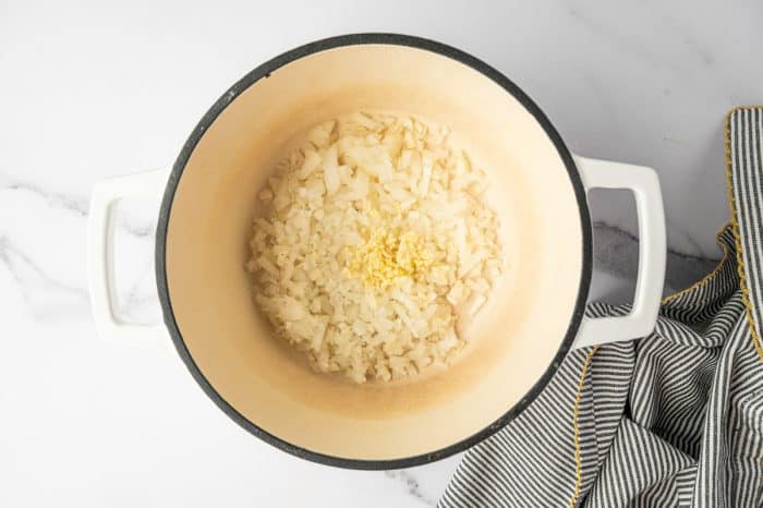 Overhead photo of finely chopped white onion and minced garlic in a cream colored enameled Dutch oven with a black rim and white handles on a light marble counter, with a black and white striped kitchen towel with yellow trim on the right.