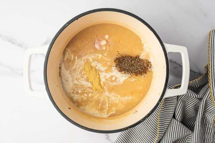 Overhead photo of a creamy soup base in a cream colored enameled Dutch oven with a black rim and white handles, showing a pile of black pepper, a pinch of salt, and a stripe of ground mustard on top with pale cream swirls, with a black and white striped towel with yellow trim on the right.