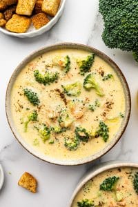Overhead photo of a beige speckled bowl filled with creamy broccoli cheddar soup topped with cracked black pepper, on a light marble surface with a white bowl of golden croutons at the top left, a large broccoli crown at the top right, and a second bowl of soup partially visible at the bottom right.