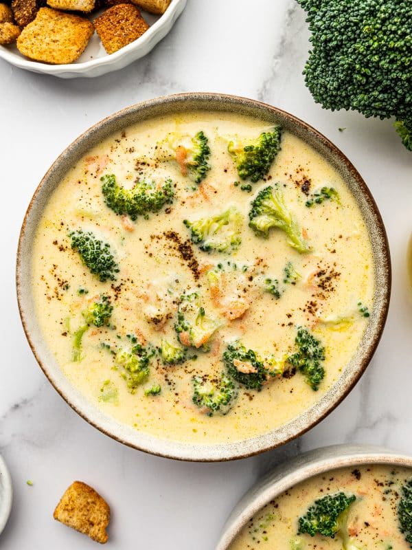 Overhead photo of a beige speckled bowl filled with creamy broccoli cheddar soup topped with cracked black pepper, on a light marble surface with a white bowl of golden croutons at the top left, a large broccoli crown at the top right, and a second bowl of soup partially visible at the bottom right.