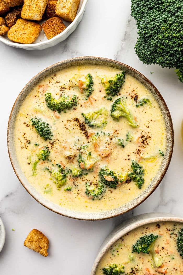 Overhead photo of a beige speckled bowl filled with creamy broccoli cheddar soup topped with cracked black pepper, on a light marble surface with a white bowl of golden croutons at the top left, a large broccoli crown at the top right, and a second bowl of soup partially visible at the bottom right.