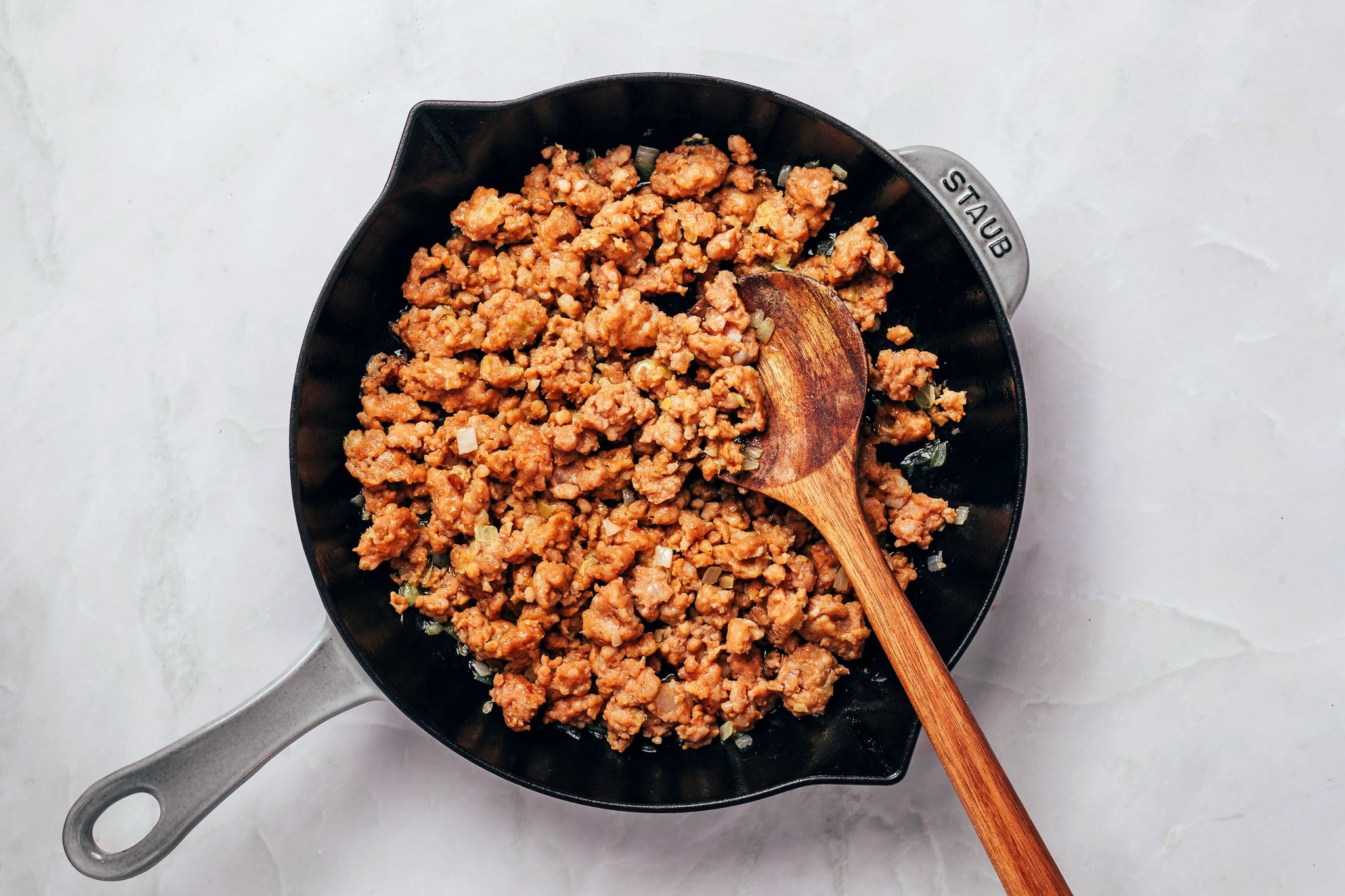 Crumbled breakfast sausage browned to a golden reddish-brown color with small pieces of softened onion in a black cast-iron skillet. A wooden cooking spoon rests among the sausage crumbles, which are spread across the bottom of the skillet.