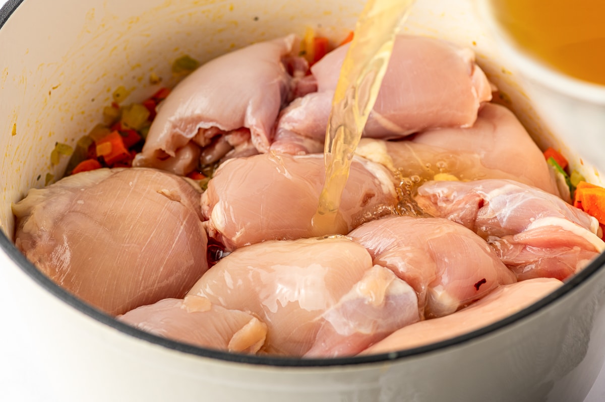 Low-sodium chicken broth being poured over boneless skinless chicken thighs and sauteed vegetables in a white Dutch oven.