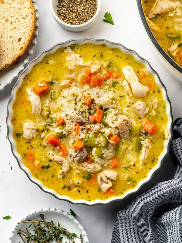 A scalloped bowl filled with chicken and rice soup containing shredded chicken, jasmine rice, carrots, celery, herbs, and black pepper, served beside rustic bread, fresh thyme, and a pot of soup.