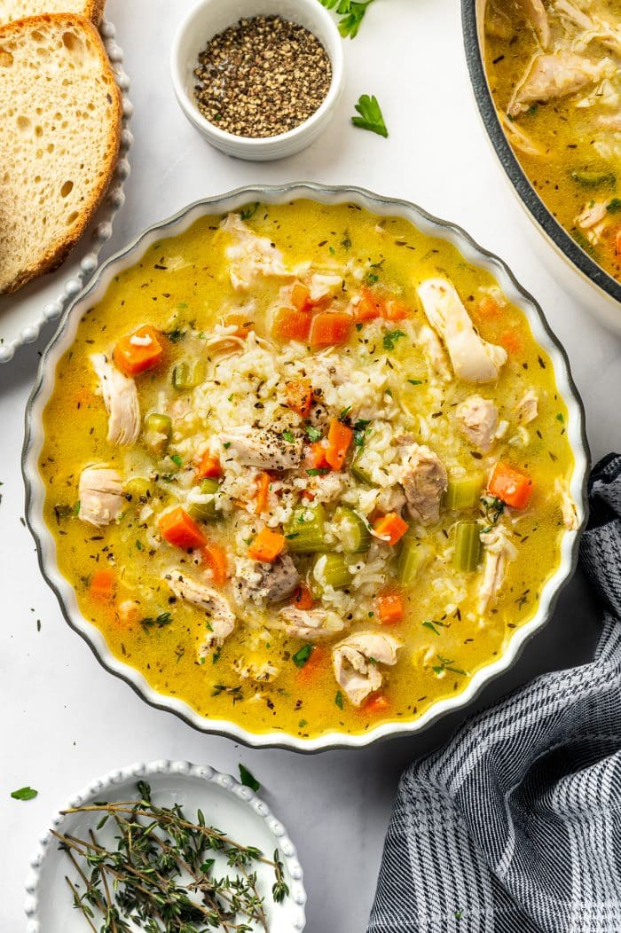A scalloped bowl filled with chicken and rice soup containing shredded chicken, jasmine rice, carrots, celery, herbs, and black pepper, served beside rustic bread, fresh thyme, and a pot of soup.