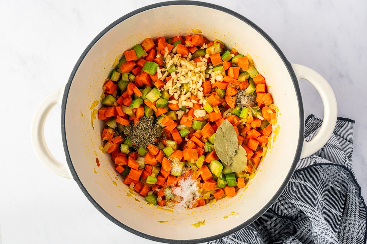 Sauteed carrots, celery, and onion topped with minced garlic, dried thyme, bay leaves, salt, and black pepper in a white Dutch oven.