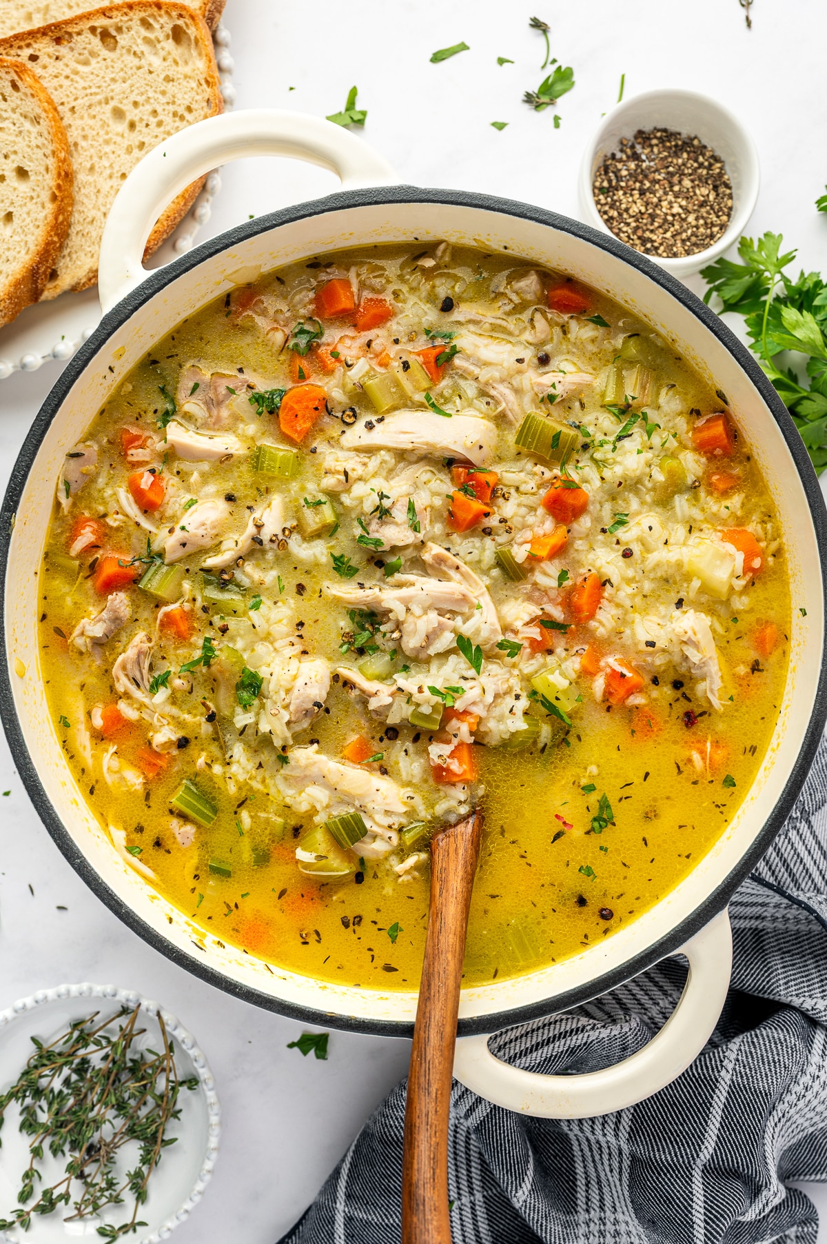 Finished chicken and rice soup in a white Dutch oven filled with shredded chicken, tender white rice, carrots, celery, and herbs in golden broth, with rustic bread, parsley, thyme, and black pepper nearby.