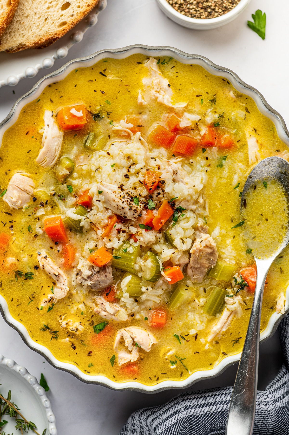 A bowl of chicken and rice soup filled with tender chicken, jasmine rice, carrots, celery, parsley, thyme, and black pepper in golden broth, with a metal soup spoon resting inside the bowl.