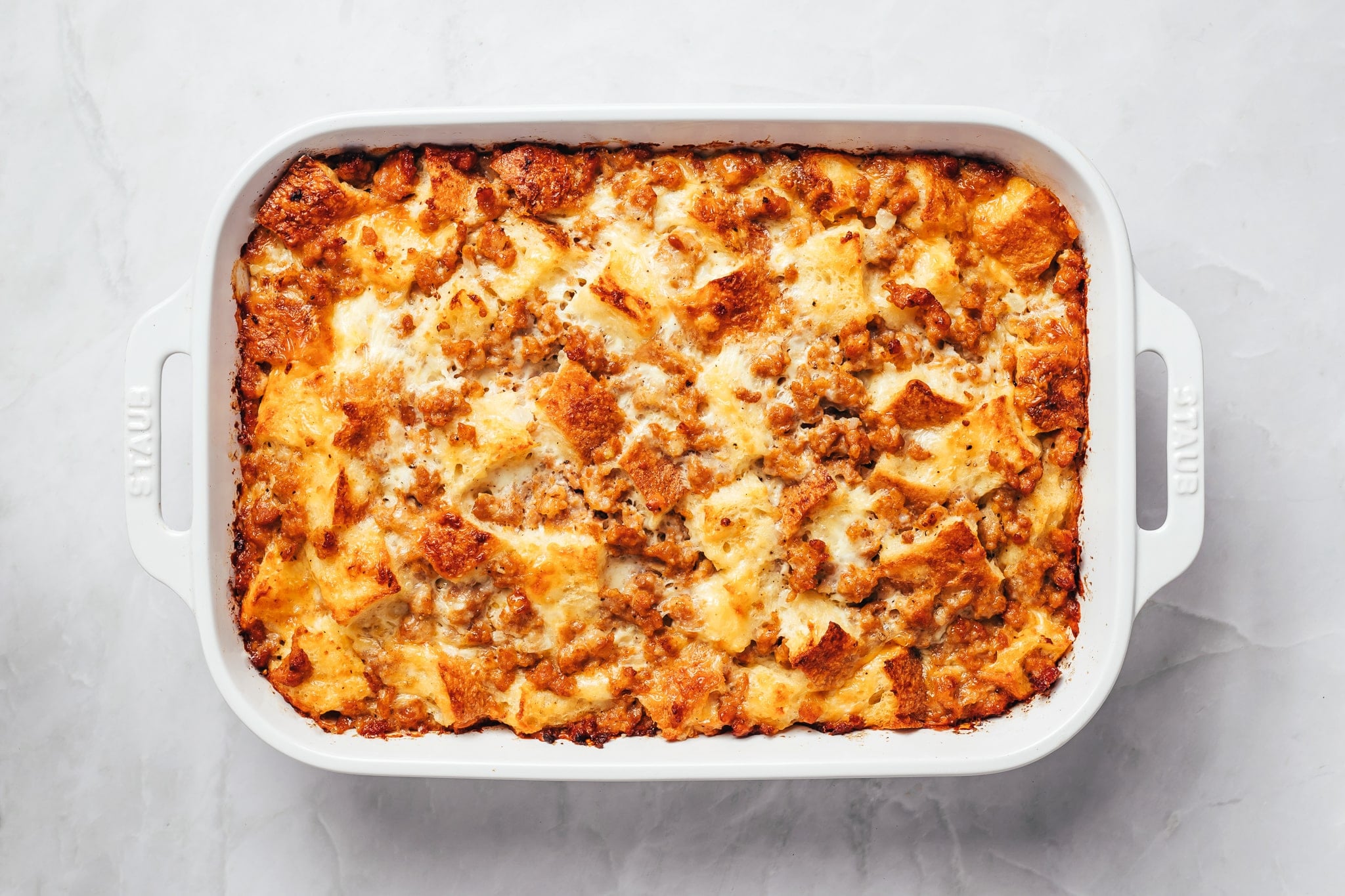 A freshly baked sausage breakfast casserole filling a white rectangular ceramic baking dish. The surface has irregular cubes of golden-brown toasted bread, melted cheese, and browned crumbles of sausage, with darker crisp edges around the dish.