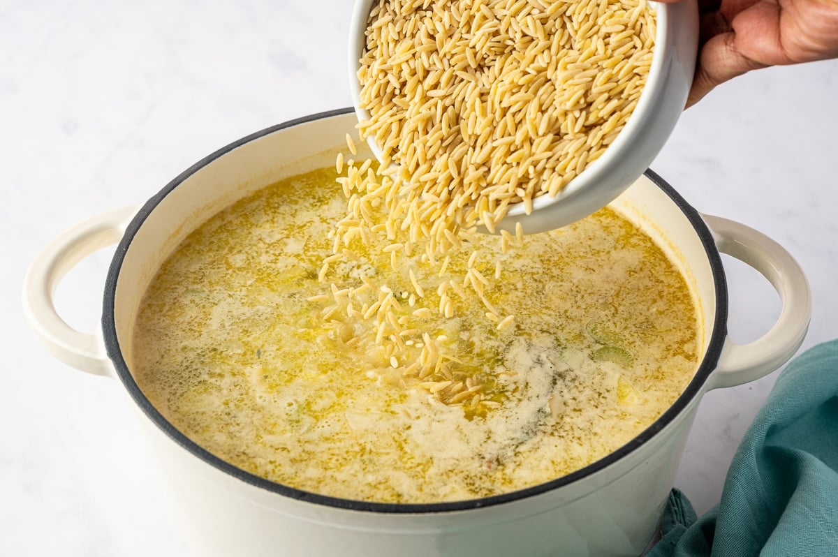 Small, uncooked, rice-shaped orzo pasta falling from a tilted white bowl into a large Dutch oven filled with pale golden chicken soup broth. A hand holds the bowl above the pot as the dry pasta pours into the liquid, with a few pieces of vegetables visible beneath the surface.