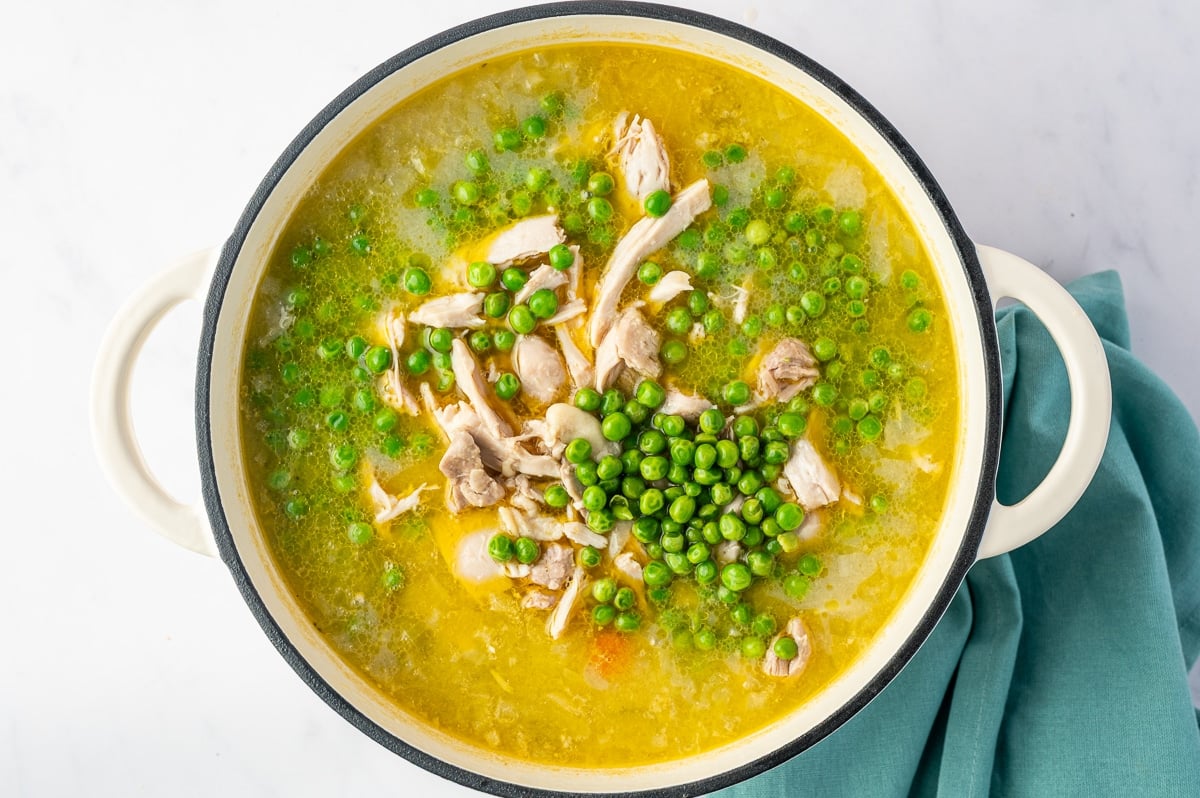 A large white Dutch oven filled with golden chicken soup broth, shredded chicken, and bright green peas. A cluster of peas sits in the center among pieces of tender chicken, while additional peas are scattered across the broth. Small pieces of carrots and celery are visible beneath the surface.