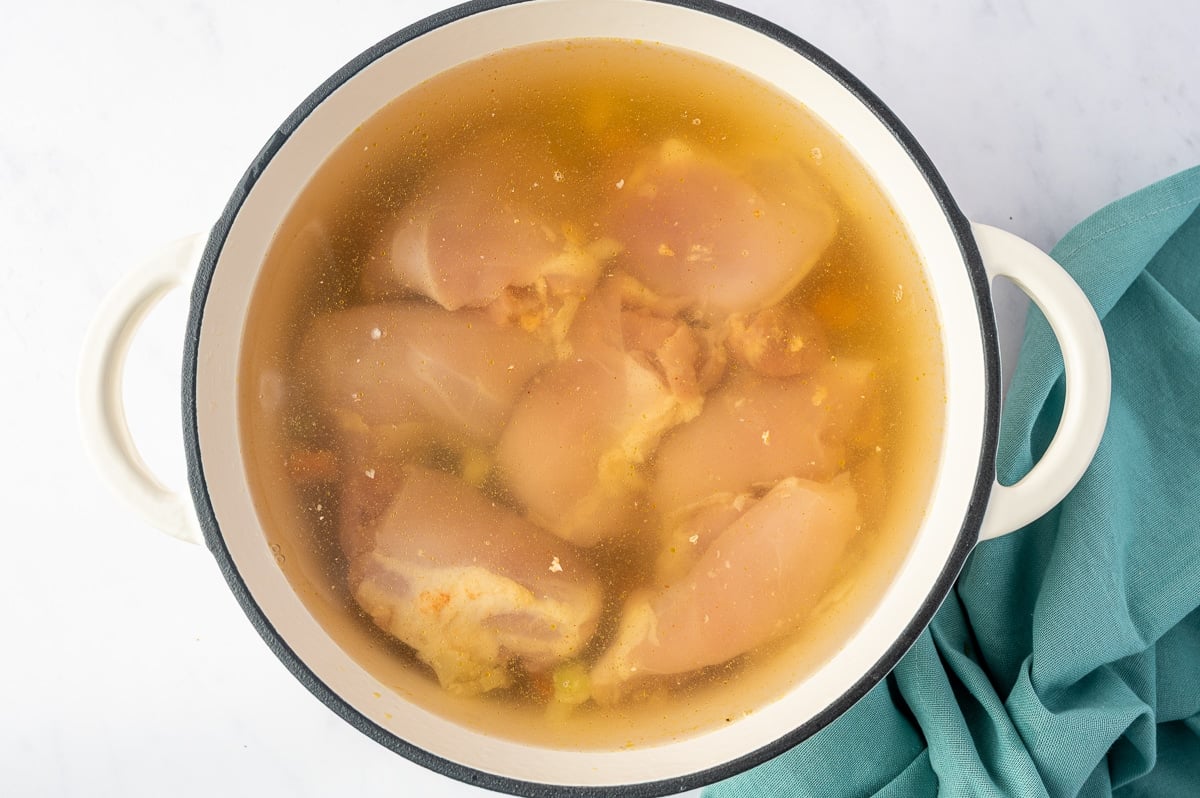 Several raw, boneless, skinless chicken thighs submerged in a large white enameled Dutch oven filled with golden chicken broth. The pink chicken pieces are visible just below the surface, with small bits of cooked vegetables and seasonings showing through the broth.