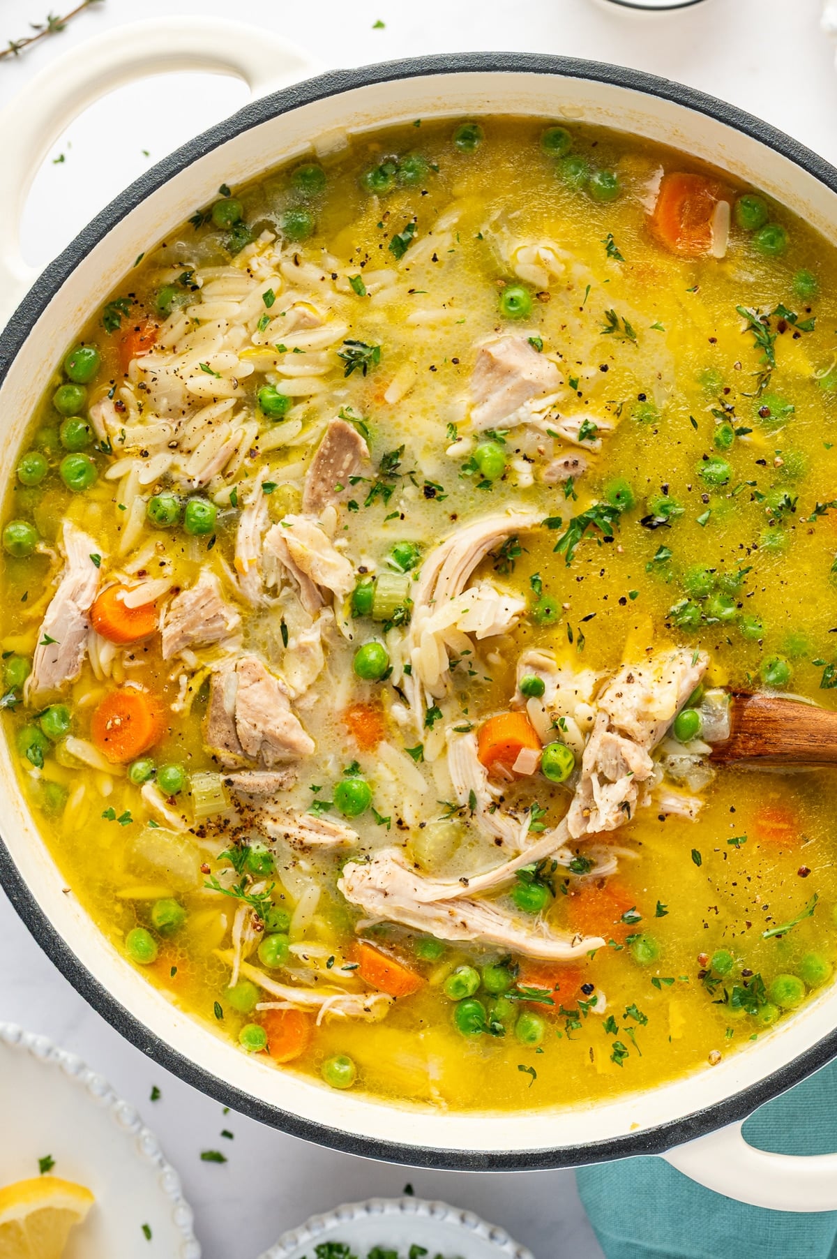 A white enameled Dutch oven filled with finished lemon chicken orzo soup. The golden broth contains generous pieces of shredded chicken, rice-shaped orzo pasta, orange carrot slices, green peas, and celery, with chopped fresh herbs and black pepper scattered over the surface. A wooden spoon rests inside the pot.