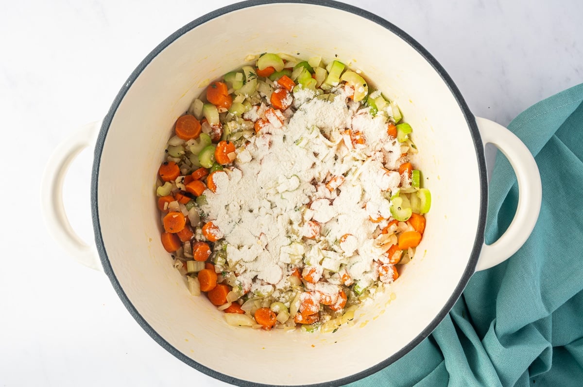 A layer of white all-purpose flour sprinkled over softened carrots, celery, onions, and herbs in a white enameled Dutch oven. The flour covers much of the center of the vegetables, while orange carrot slices and green celery pieces remain visible around the edges.