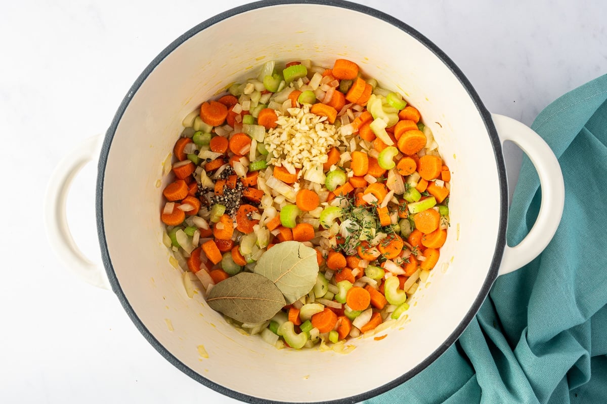 Sautéed carrots, onions, and celery in a white Dutch oven topped with a small mound of finely minced garlic, cracked black pepper, fresh thyme leaves, and two whole dried bay leaves. The seasonings are still separated on top of the colorful vegetables, ready to be stirred together.
