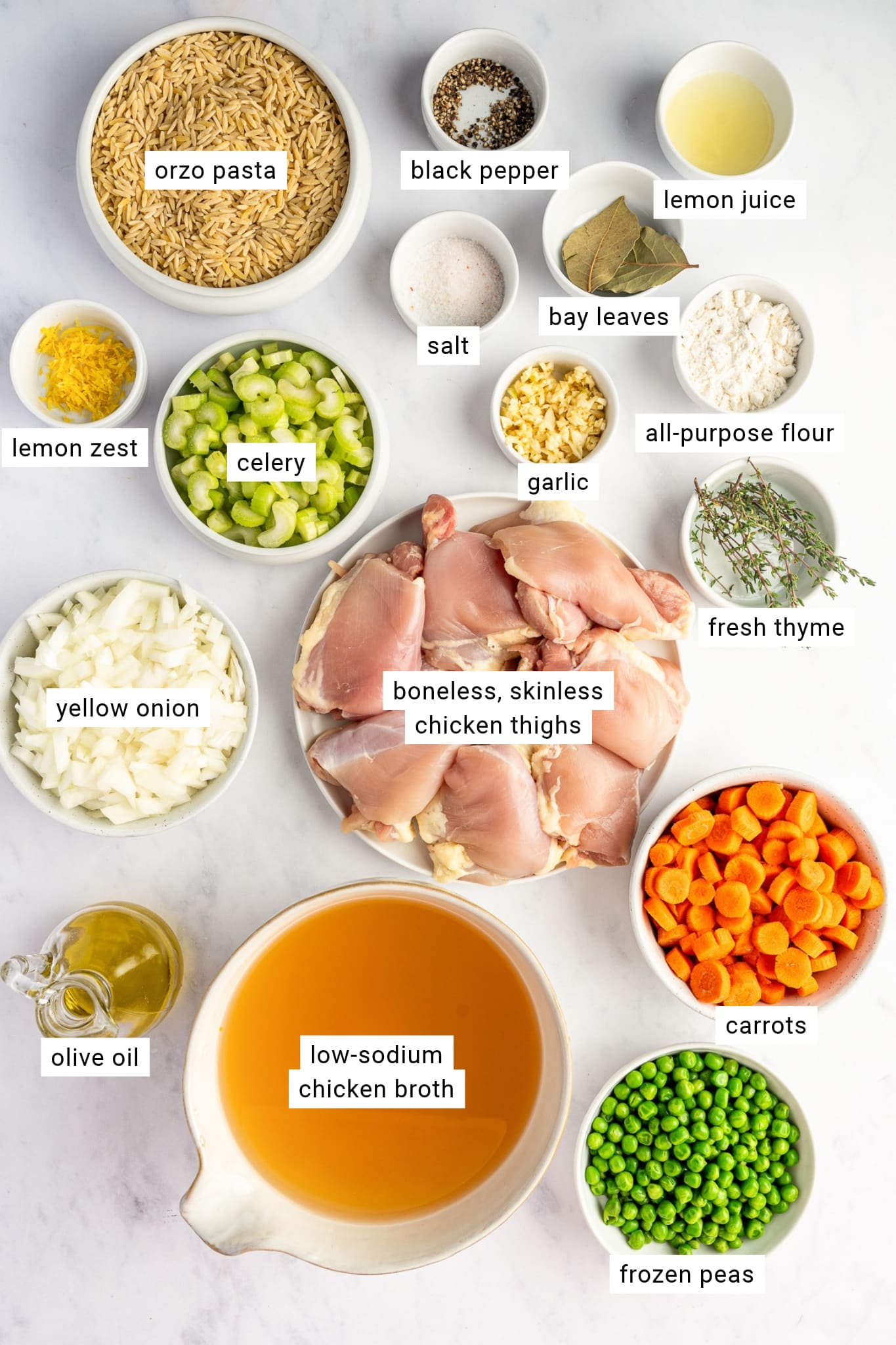 Ingredients for lemon chicken orzo soup arranged in separate bowls with labels, including raw boneless chicken thighs, uncooked orzo pasta, chopped carrots, celery, yellow onion, frozen peas, chicken broth, olive oil, fresh lemon juice and zest, garlic, thyme, bay leaves, flour, salt, and black pepper.