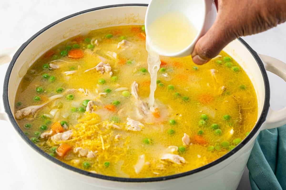 A hand pouring pale yellow lemon juice from a small white bowl into a large Dutch oven filled with chicken orzo soup. Fresh yellow lemon zest is visible on the surface of the golden broth, along with pieces of shredded chicken, orange carrots, green peas, and orzo pasta.