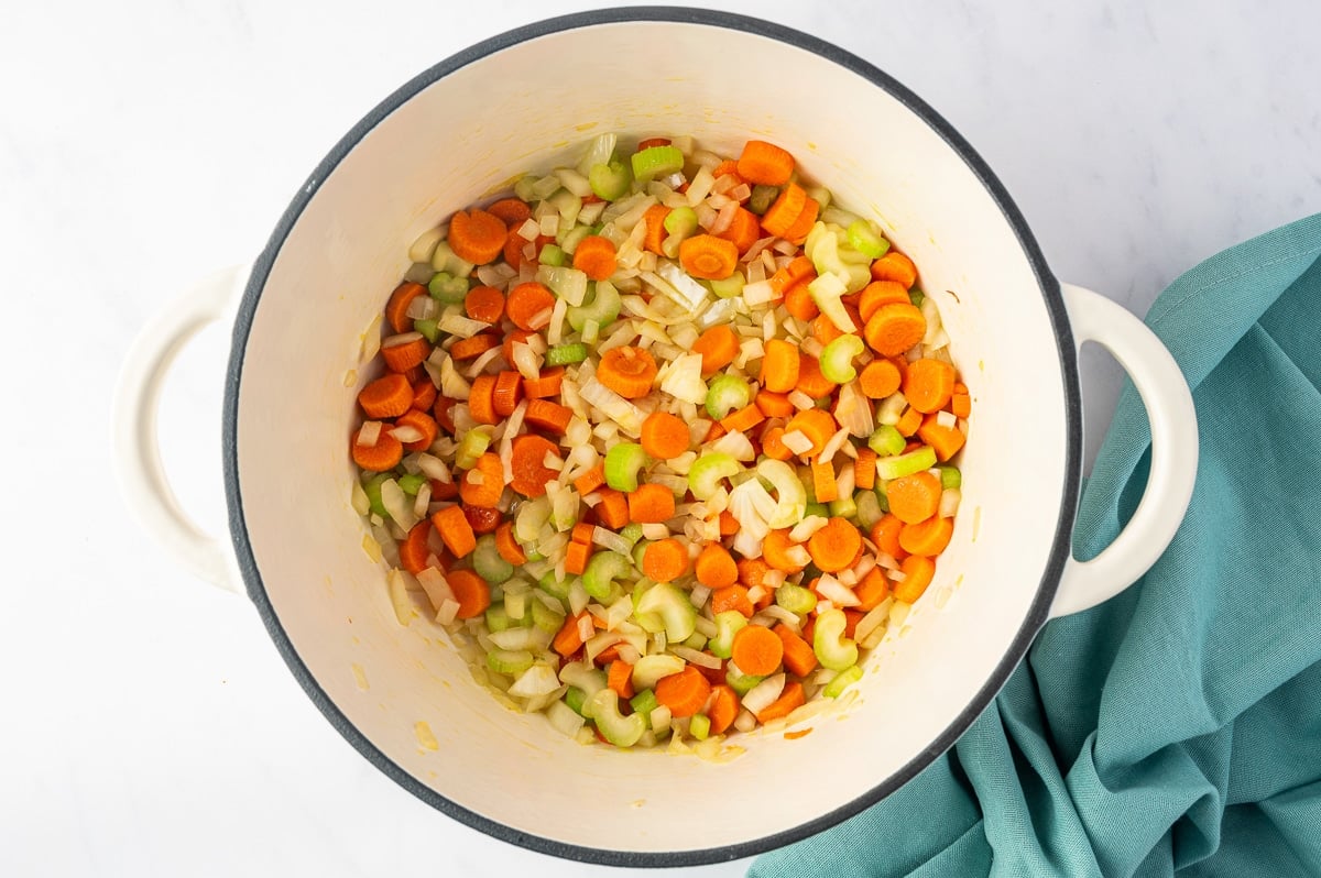 Chopped carrots, diced yellow onions, and sliced celery mixed together in a large white enameled Dutch oven. The orange carrots, pale translucent onions, and bright green celery are lightly coated in oil and beginning to soften, with a folded teal kitchen towel beside the pot.