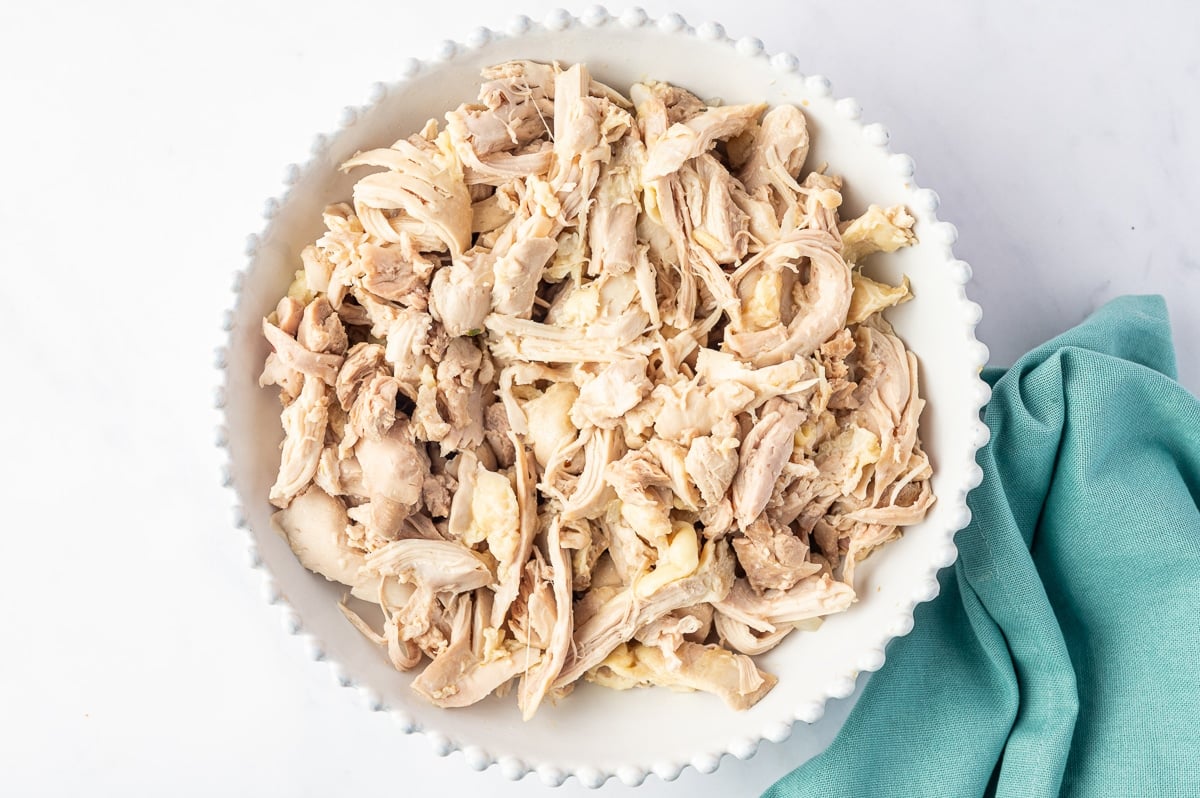 A white scalloped bowl filled with cooked chicken thighs that have been shredded into irregular strips and bite-sized pieces. The tender, pale chicken has a mixture of thin shredded strands and larger chunks, with a folded teal kitchen towel beside the bowl.