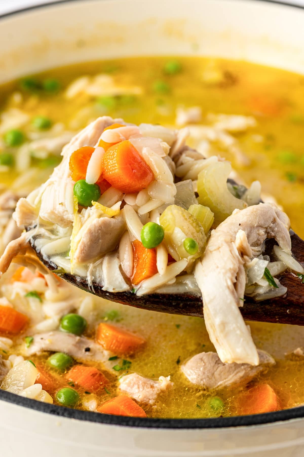 A dark wooden spoon holding a generous serving of lemon chicken orzo soup above a pot of golden broth. The spoon is piled with shredded chicken, small rice-shaped orzo pasta, orange carrot pieces, pale green celery, and bright green peas. A long strip of chicken hangs over the spoon's edge, with more soup and vegetables visible beneath it.