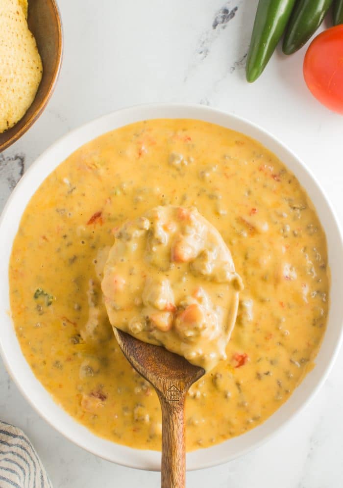 Wooden spoon filled with rotel dip resting on top of a white bowl filled with cheesy rotel dip.