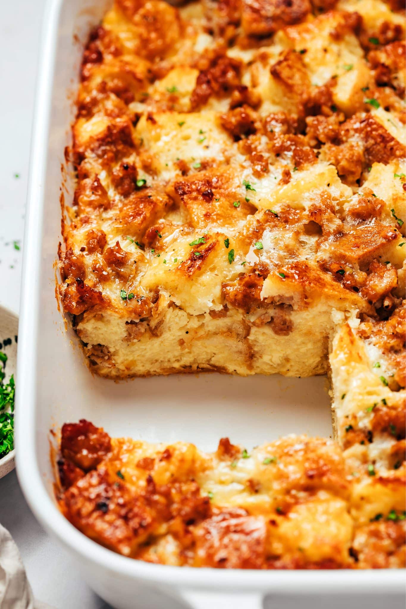 A white ceramic baking dish containing sausage breakfast casserole with a square portion removed. The cut edge reveals a thick, pale golden layer of baked egg custard and softened sourdough bread beneath a browned topping of sausage, melted cheese, toasted bread, and small flecks of green parsley.