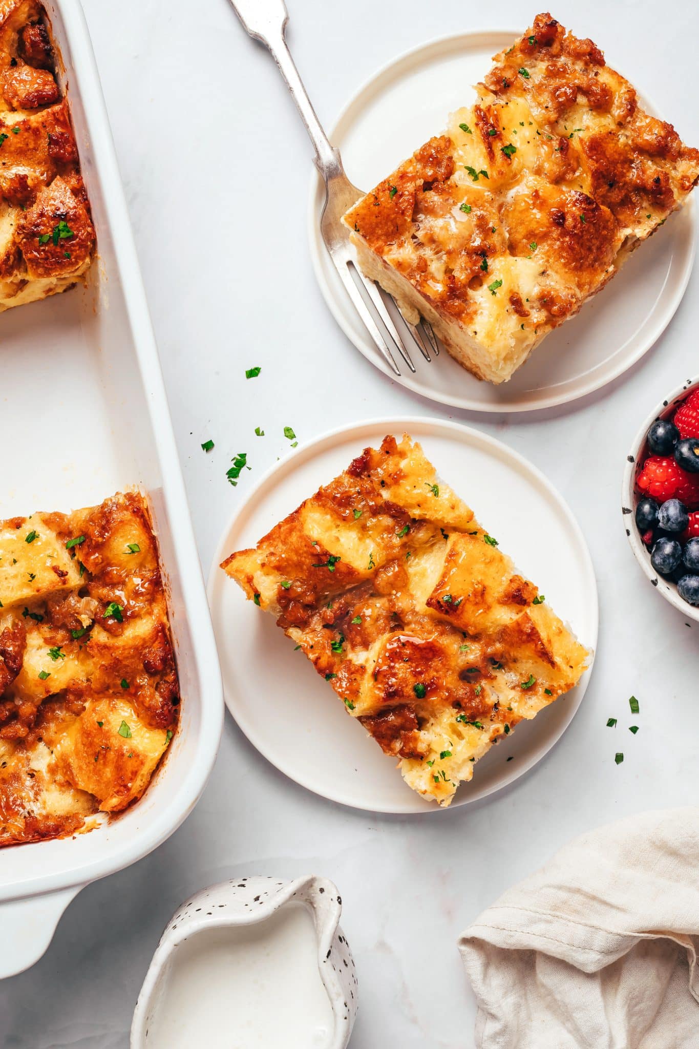 Two square servings of golden-brown sausage breakfast casserole arranged on separate white plates, each topped with melted cheese, browned sausage, toasted sourdough bread, and small flecks of parsley. Part of the remaining casserole is visible in a white baking dish, with a bowl of raspberries and blueberries, a small cream pitcher, a fork, and a light-colored linen napkin nearby.