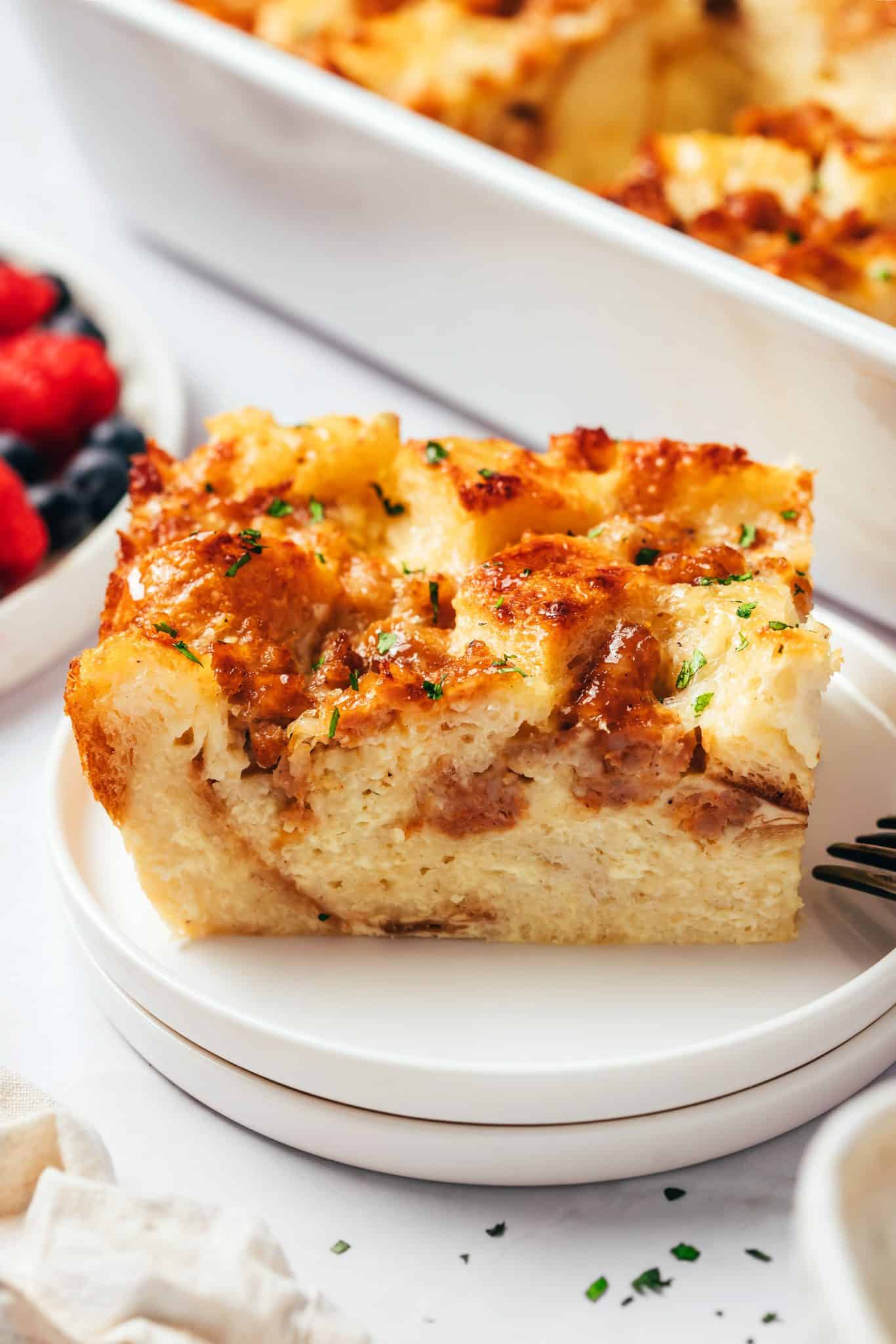 A thick rectangular portion of sausage breakfast casserole served on a round white plate. The slice has a golden-brown surface with toasted sourdough bread, melted cheese, small pieces of browned sausage, and chopped parsley. The interior shows soft layers of baked egg custard and bread, with a white baking dish and fresh berries visible behind the plate.