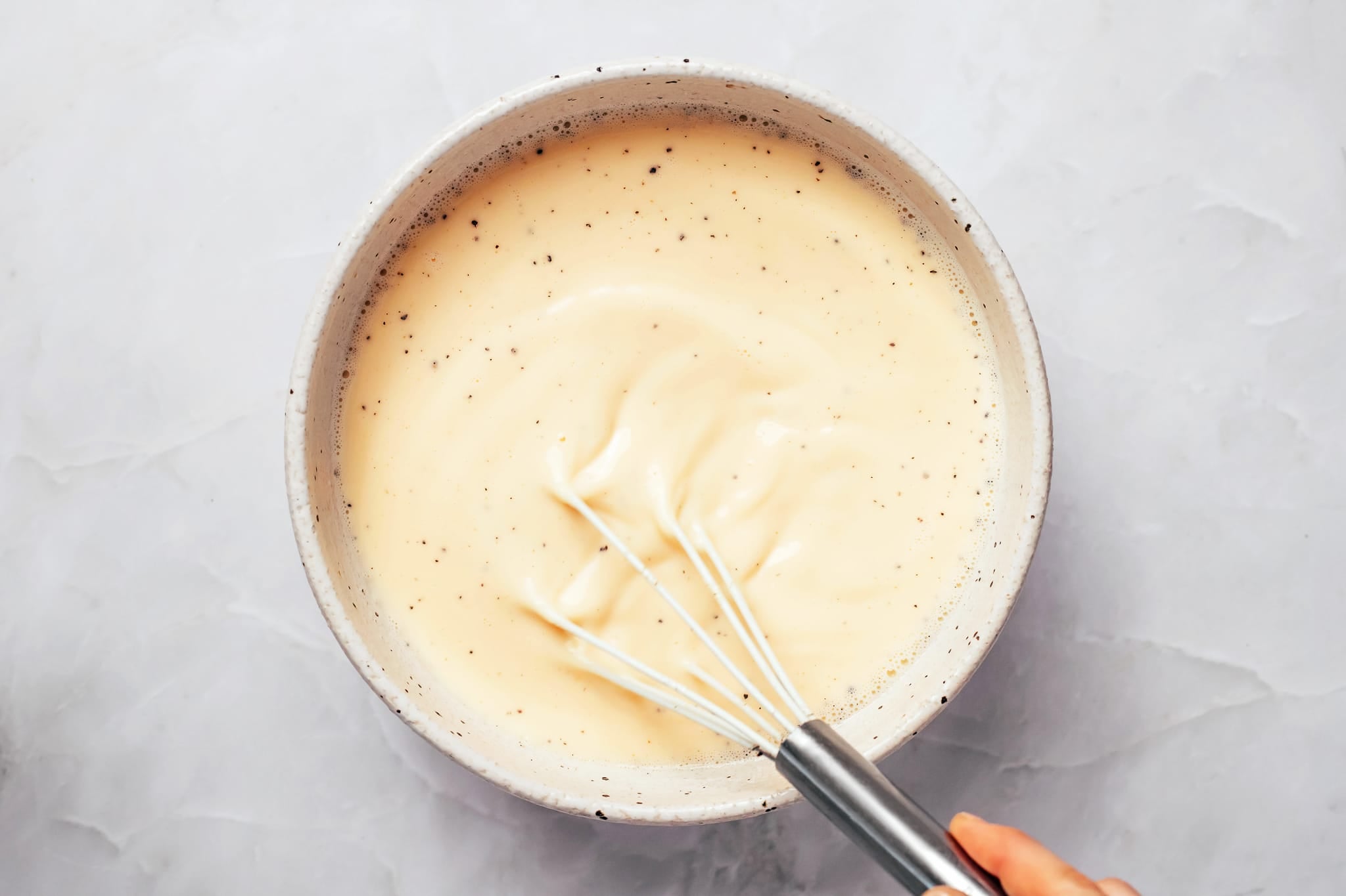 A round, cream-colored ceramic mixing bowl filled with a pale yellow mixture of beaten eggs, milk, and small dark flecks of seasoning. A metal whisk is partially submerged in the smooth liquid, with small bubbles visible around the edges of the bowl.