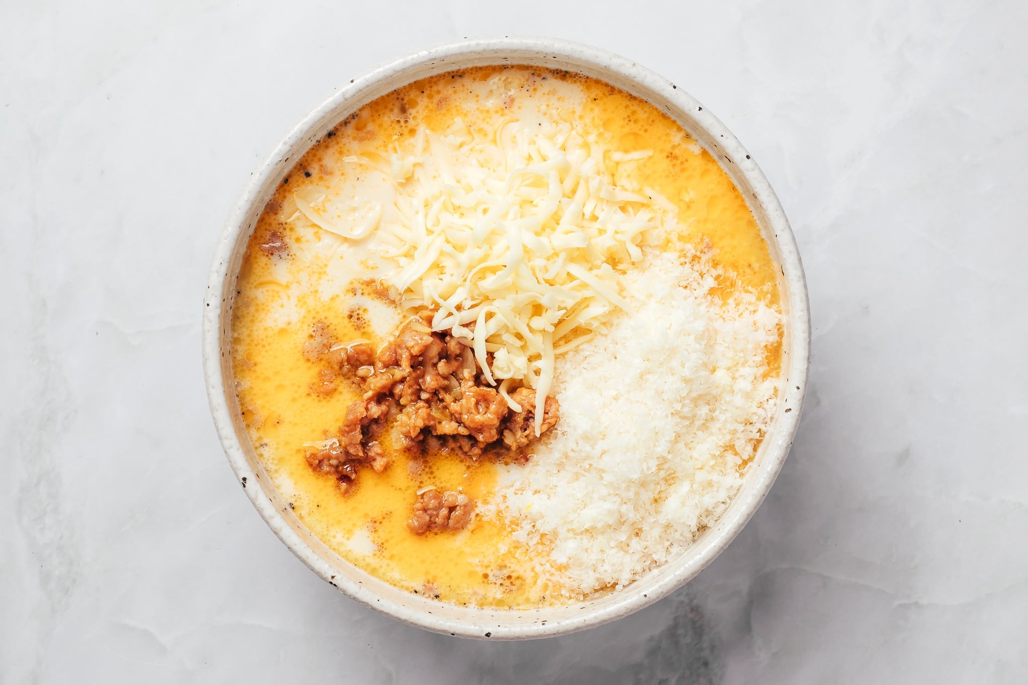 A large ceramic mixing bowl containing a pale orange egg and milk mixture topped with separate portions of browned crumbled sausage, shredded pale yellow cheese, and finely grated white Parmesan cheese. The ingredients have been added to the bowl but are not yet fully mixed together.
