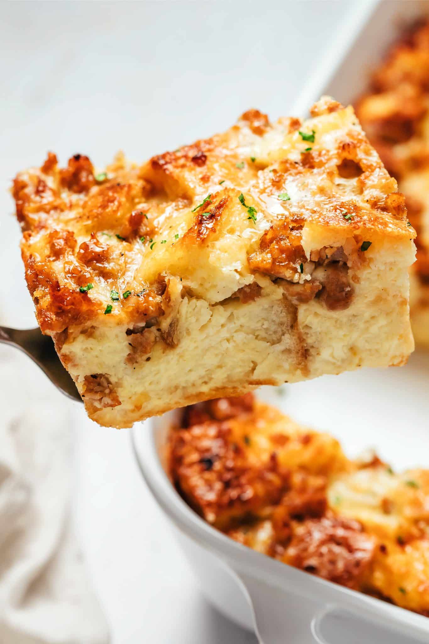 A thick square slice of sausage breakfast casserole resting on a metal serving spatula, with the remaining casserole visible in a white ceramic baking dish behind it. The slice has a golden-brown top with melted cheese, browned sausage pieces, toasted sourdough bread, and small green parsley flecks. The interior is pale golden, soft, and thick with baked bread and egg custard.