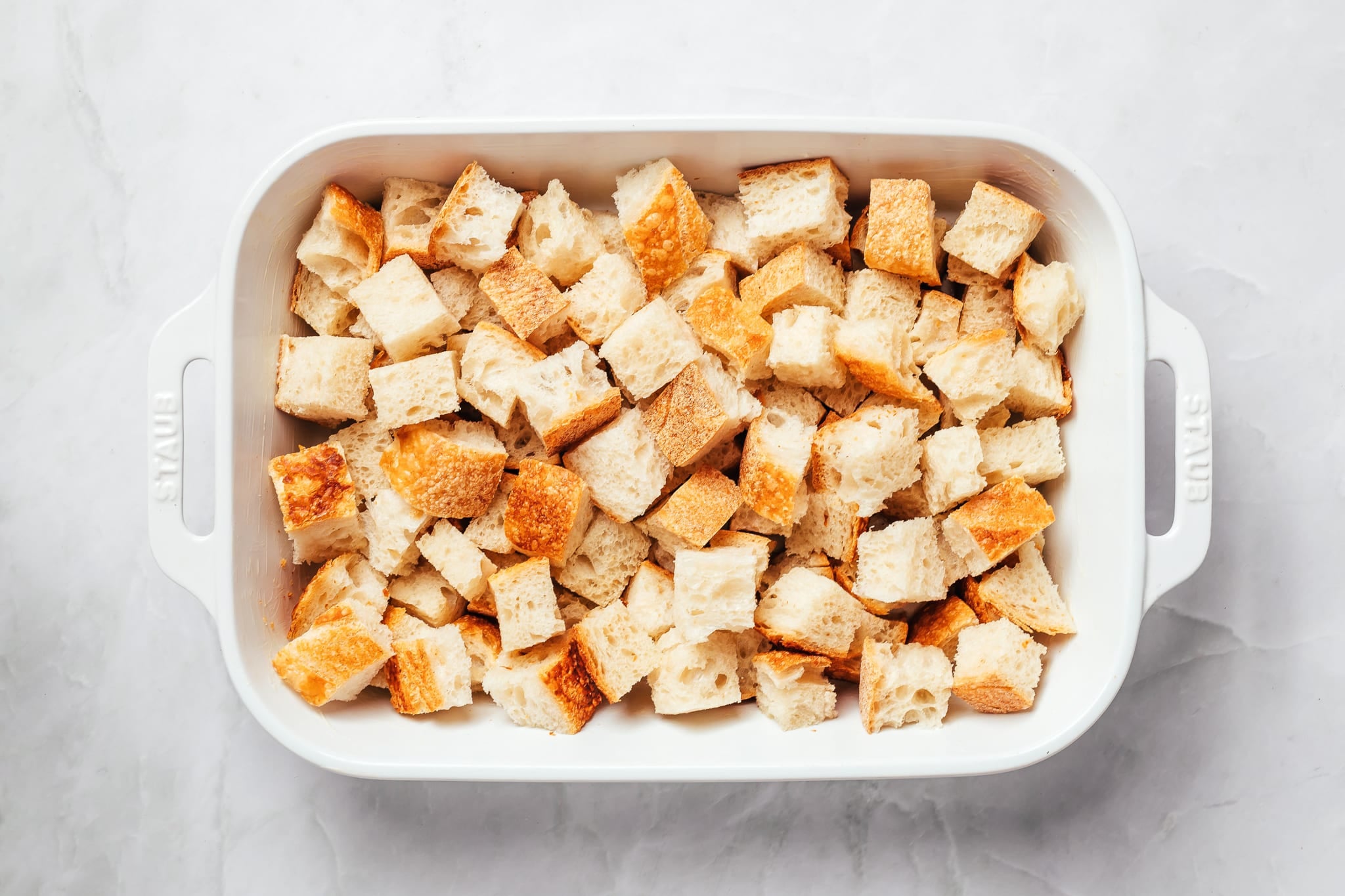 A white rectangular ceramic baking dish filled with large, uneven cubes of sourdough bread. The bread has airy white interiors and golden crusts, with the pieces loosely piled together and covering the entire bottom of the dish.