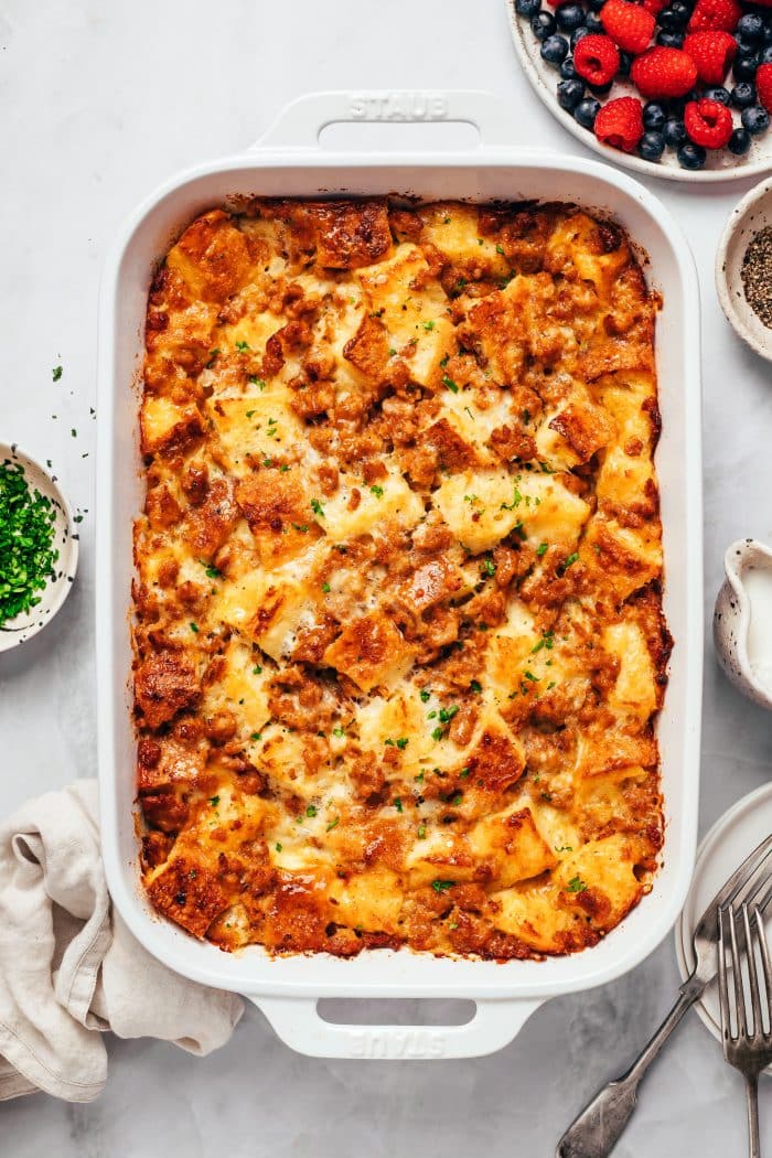 A whole sausage breakfast casserole in a white rectangular baking dish, with a golden, uneven surface of toasted sourdough bread, browned sausage, and melted cheese. Small pieces of chopped green parsley are scattered over the top. A bowl of raspberries and blueberries, small serving bowls, and silverware sit around the casserole.