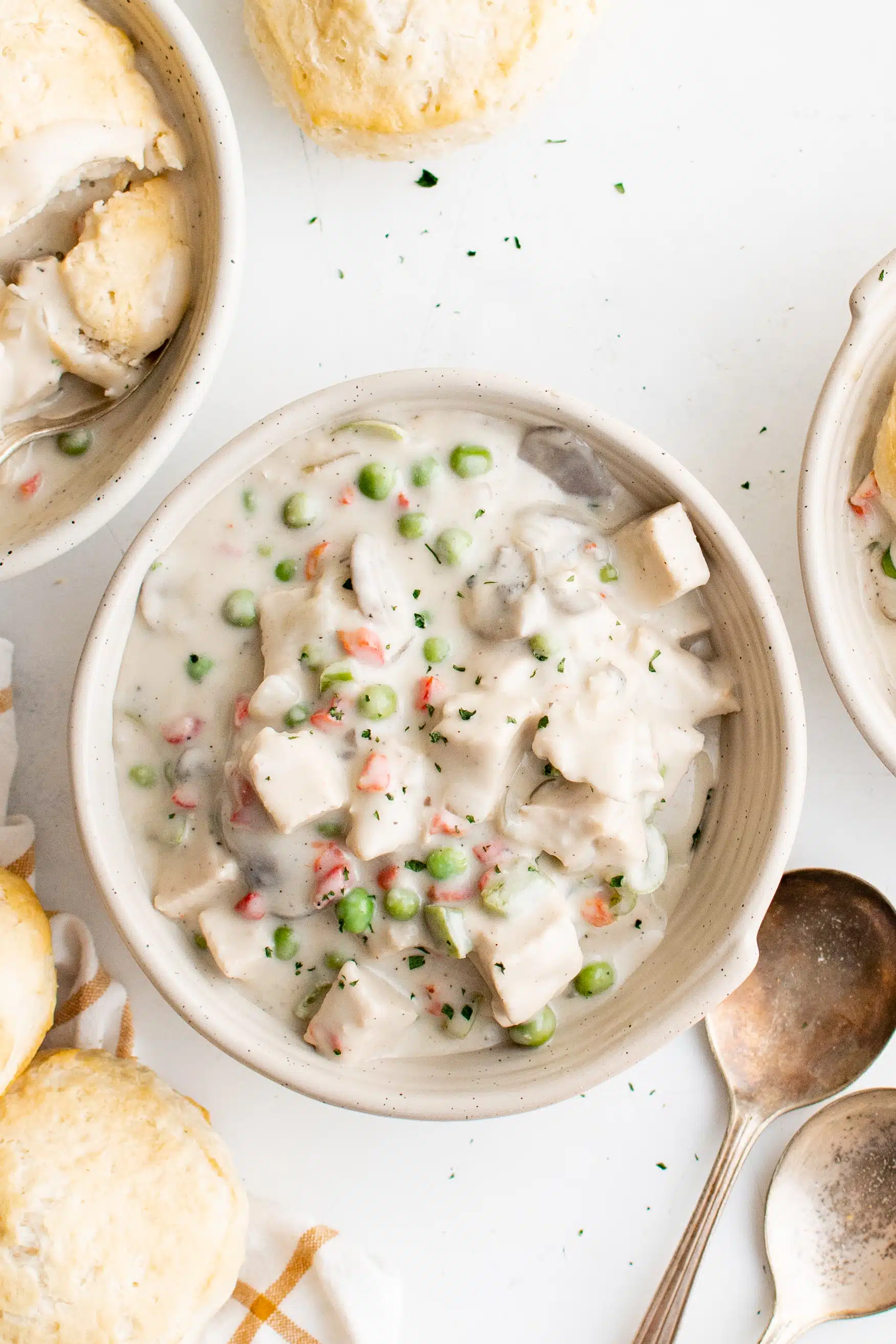 White soup bowl filled with creamy chicken a la king topped with flakey biscuit and surrounded by additional biscuits and a spoon.