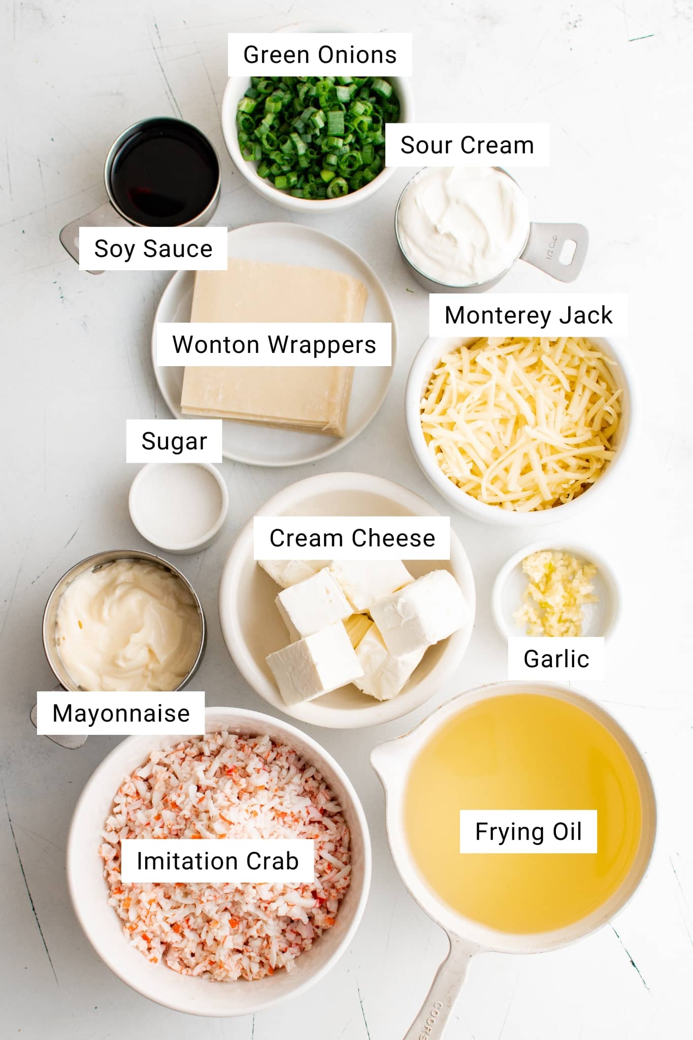 Overhead view of neatly arranged ingredients for crab rangoon dip on a white surface, each labeled with text. The spread includes a bowl of chopped green onions, sour cream in a measuring cup, shredded Monterey Jack cheese, minced garlic, cubed cream cheese, a small bowl of sugar, mayonnaise, shredded imitation crab meat, soy sauce in a measuring cup, a stack of wonton wrappers, and a measuring cup filled with frying oil.