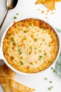 Golden baked crab rangoon dip in a white round baking dish, topped with melted cheese and sliced green onions, surrounded by crispy wonton chips on a white table.