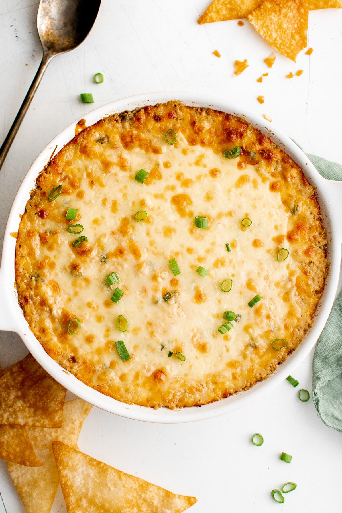Golden baked crab rangoon dip in a white round baking dish, topped with melted cheese and sliced green onions, surrounded by crispy wonton chips on a white table.
