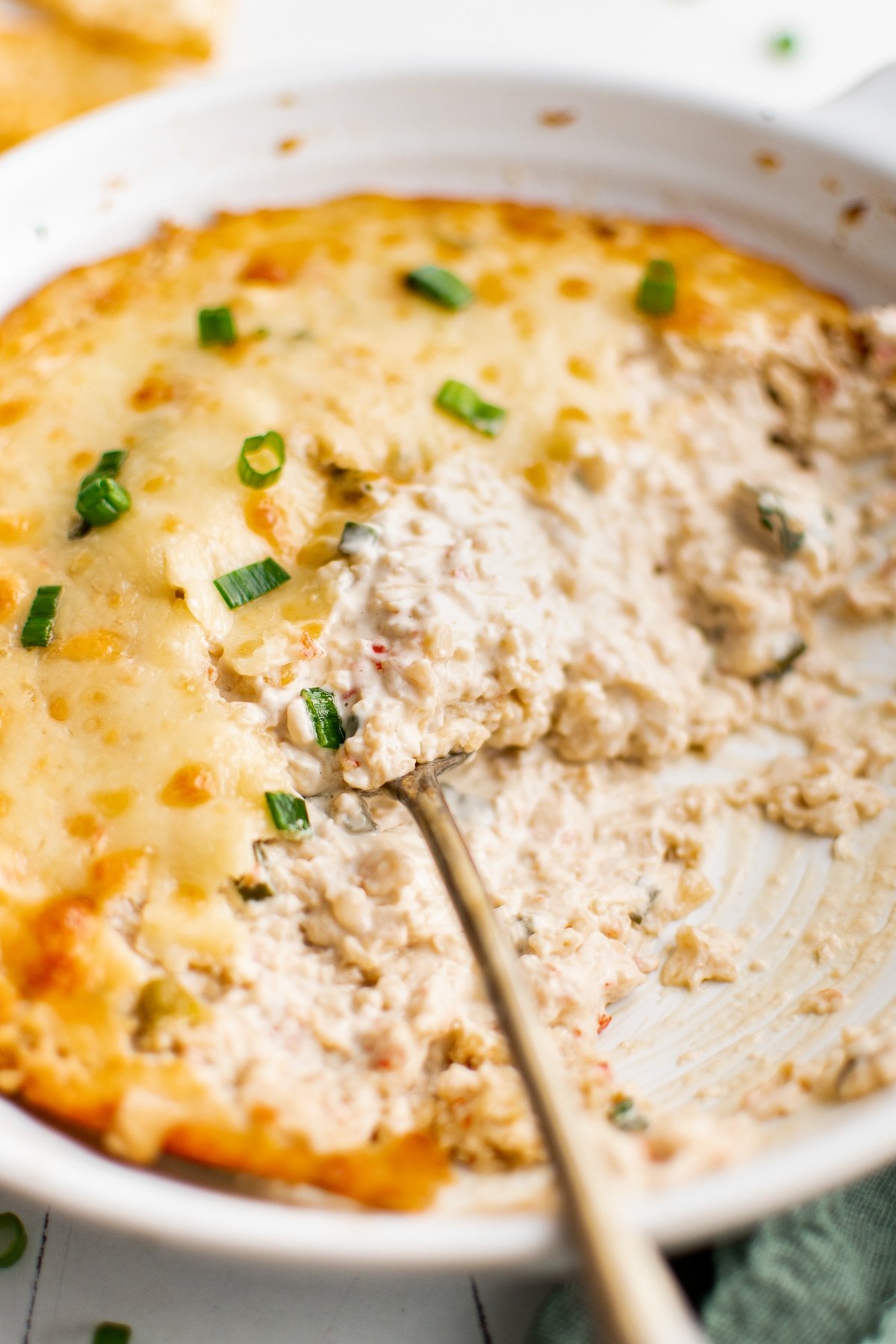 A spoon scooping into a baking dish filled with hot, cheesy crab rangoon dip, showing creamy filling beneath the melted golden cheese topping.