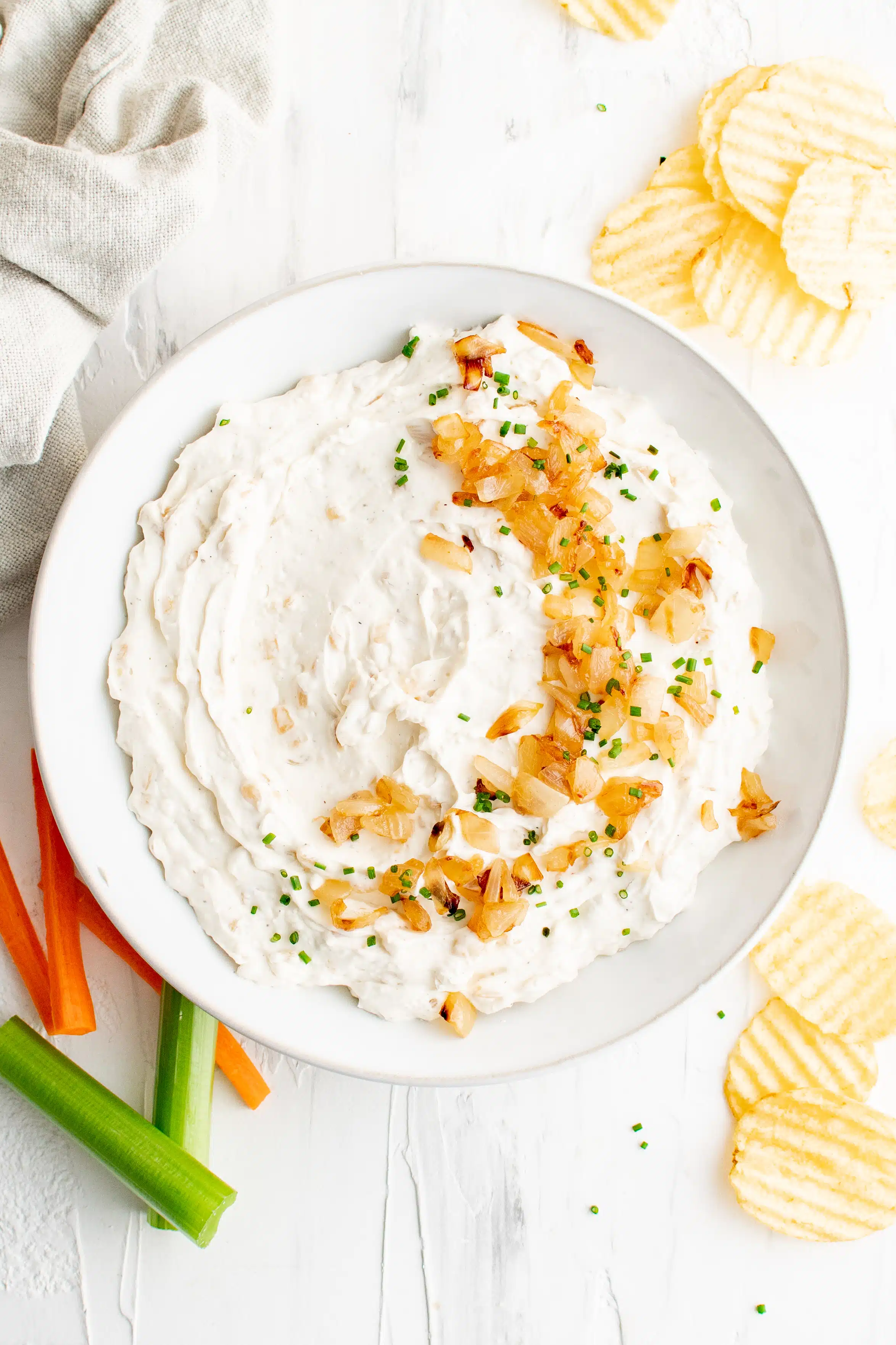 Large round white bowl filled with homemade French onion dip topped with golden caramelized onions and chopped chives on a table with potato chips and carrot and celery sticks.