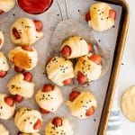 Baking sheet filled with baked mini pigs in a blanket served with ketchup and dijon mustard.