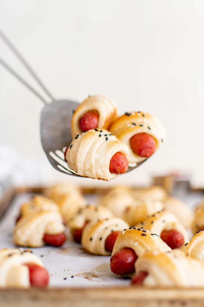 Large metal serving spoon holding three small pigs in a blanket hovering over a baking sheet filled with more mini pigs in a blanket.