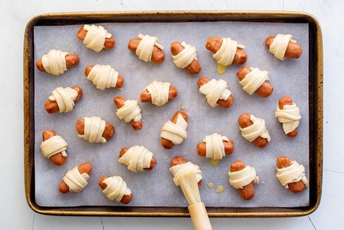 Crescent-wrapped sausages arranged on a baking sheet while being brushed with egg wash.